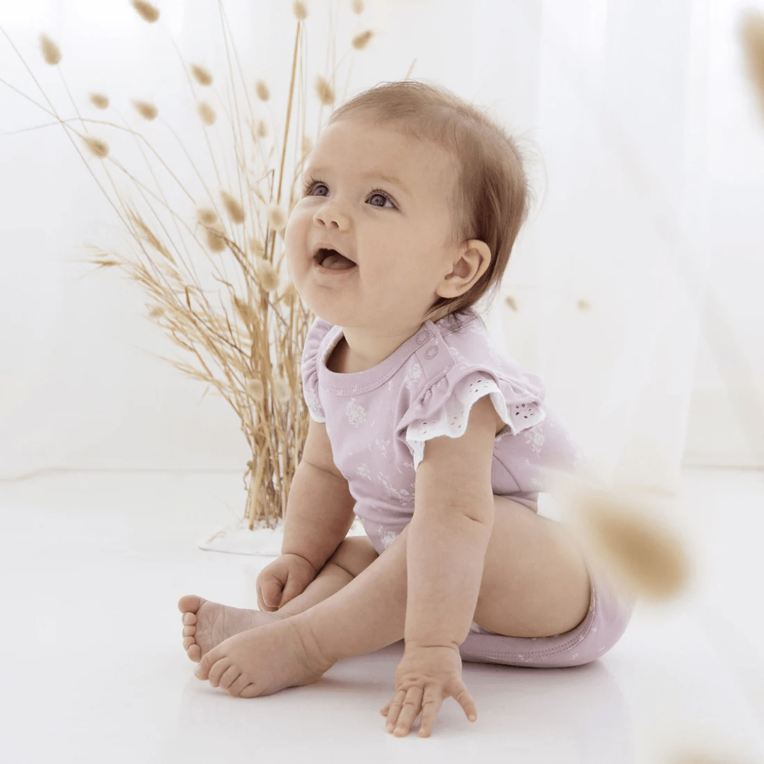 A baby is sitting on the floor in front of a white background wearing an Aster & Oak Organic Cotton Willow Floral Flutter Onesie - LUCKY LAST - NEWBORN ONLY.