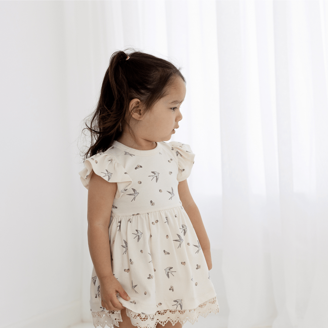 Little girl standing in front of a sheer white curtain wearing an off-white swallow-print dress with ruffle sleeves and delicate lace trim