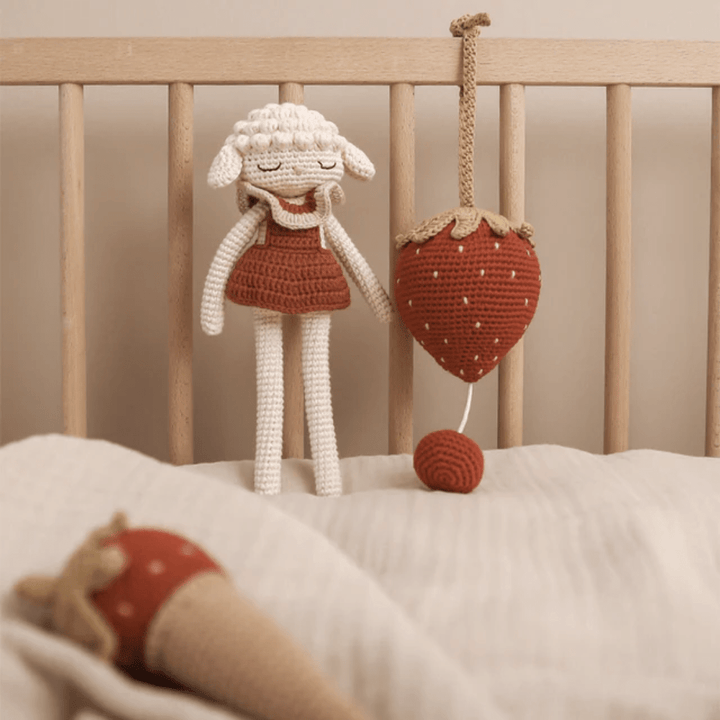 An Patti Oslo Organic Cotton Luna Lamb crafted using organic cotton is carefully placed alongside a strawberry toy in a crib.
