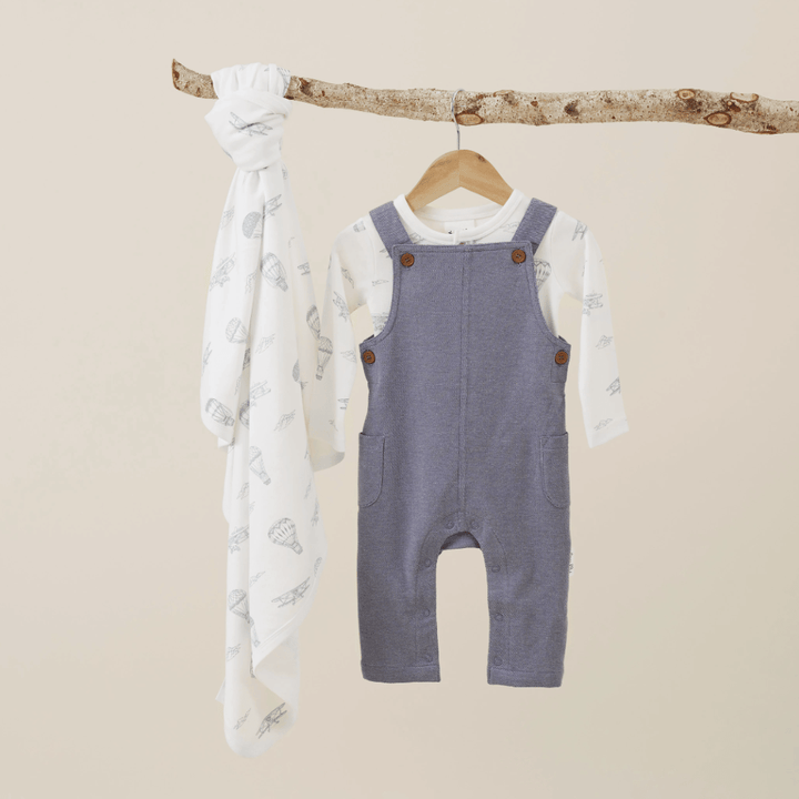Aster & Oak Organic Dark Chambray Overalls, including adjustable sizing, hanging on a wooden branch against a beige background.