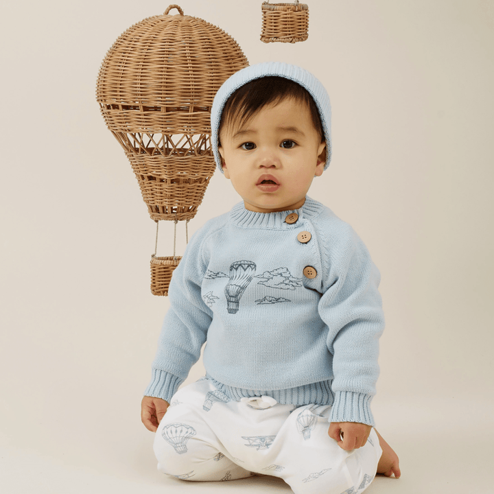 Toddler in a blue sweater and Aster & Oak organic harem pants sitting in front of a wicker hot air balloon decoration.