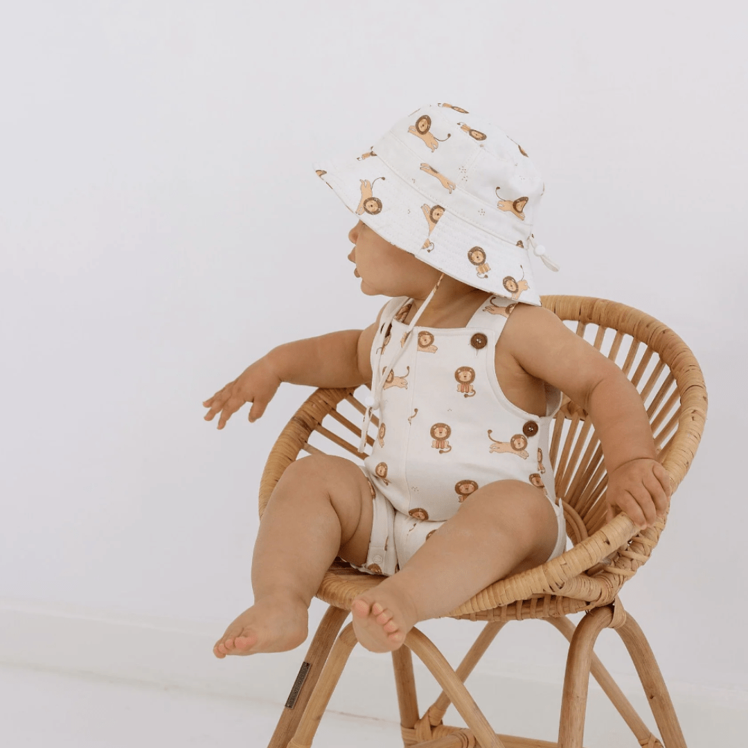 A baby sitting in a wicker chair wearing an Aster & Oak Bucket Hat - LUCKY LASTS - MEADOW & LION ONLY with adjustable straps.