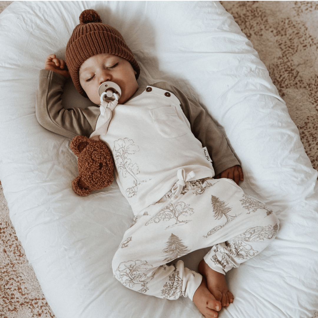 A sleeping infant with a pacifier, wearing Aster & Oak Organic Harem Pants made from GOTS-certified organic cotton, and lying next to a teddy bear on a white background.