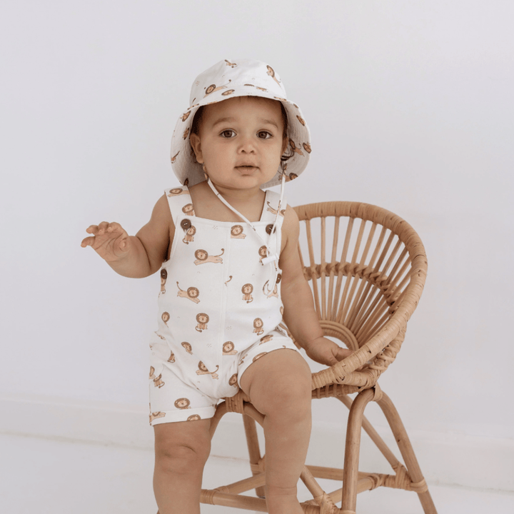 A baby sitting on a wicker chair wearing an Aster & Oak Bucket Hat - LUCKY LASTS - MEADOW & LION ONLY romper.