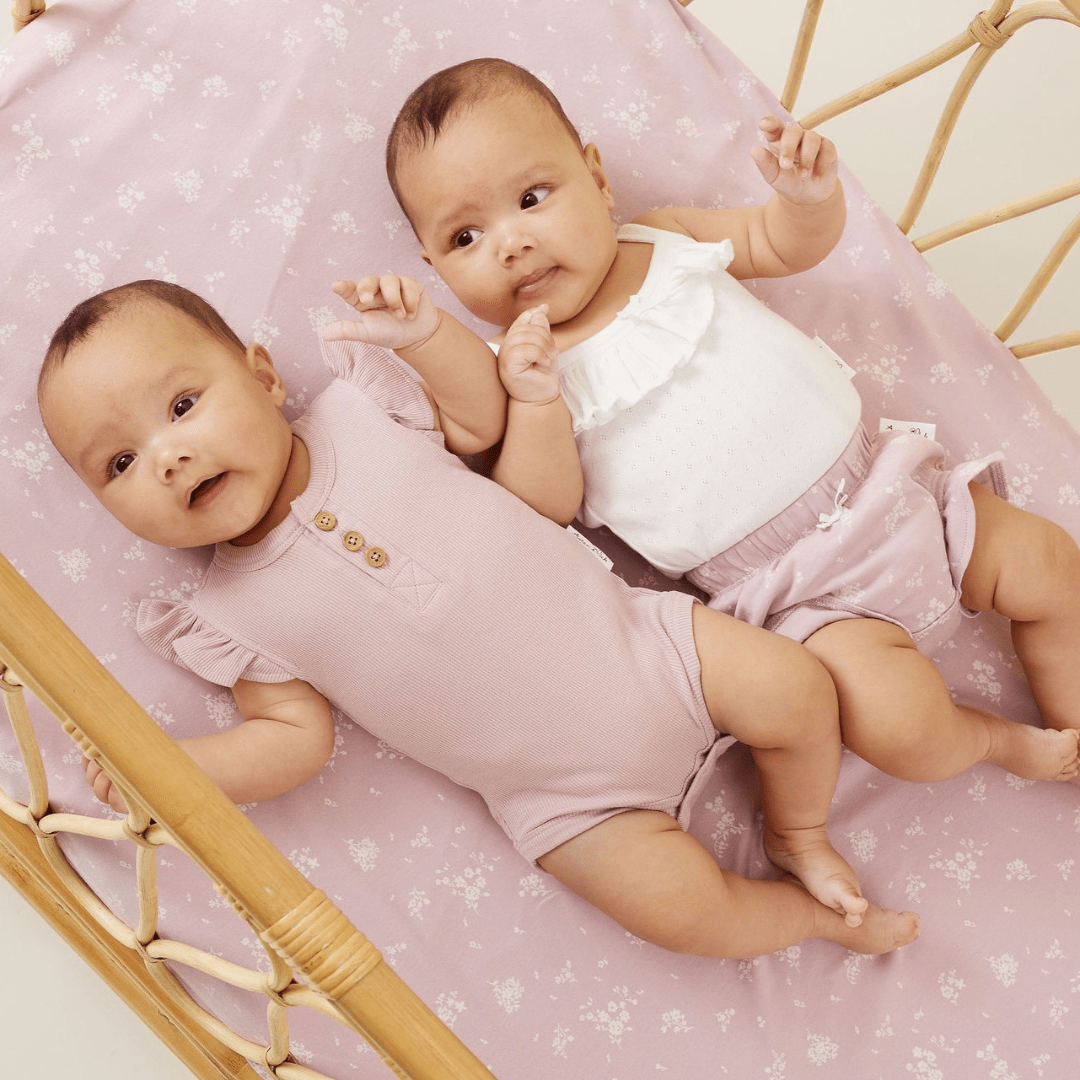 Two babies wearing Aster & Oak Organic Cotton Dawn Pink Rib Flutter Onesies, laying on a pink blanket in a crib.
