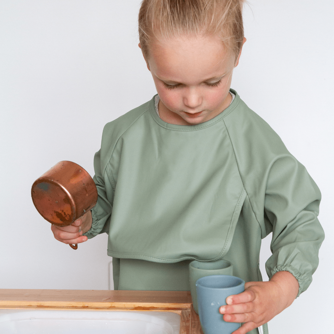 A little girl wearing a green apron and holding a cup, protected by a Zazi Recycled Full-Sleeved Bib (Multiple Variants).