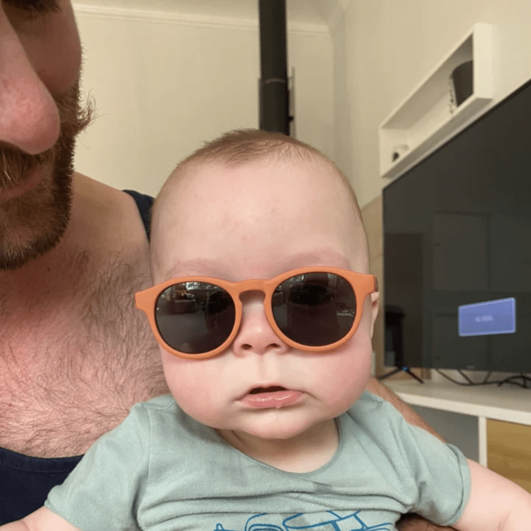 A baby wearing Zazi Shades Baby & Toddler Sunglasses in front of a tv, with UV400 protection.