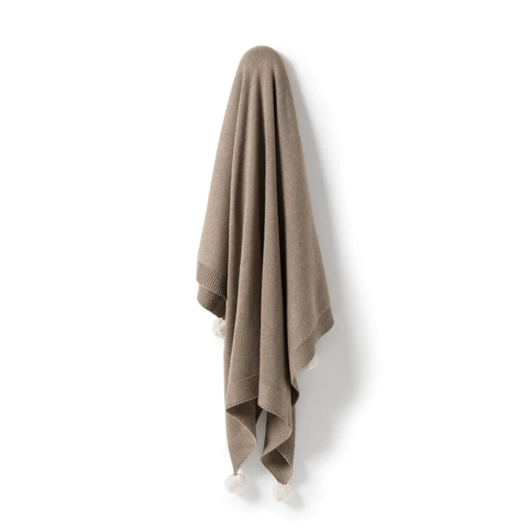 A Wilson & Frenchy Knitted Baby Blanket in beige cotton draped over a hook, casting a soft shadow on a light wall.