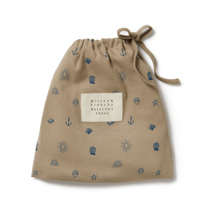 Fabric drawstring bag with product label for a Wilson & Frenchy Organic Rib Bassinet Sheet in LUCKY LASTS, featuring a pattern of blue anchors and wheels on a beige background.