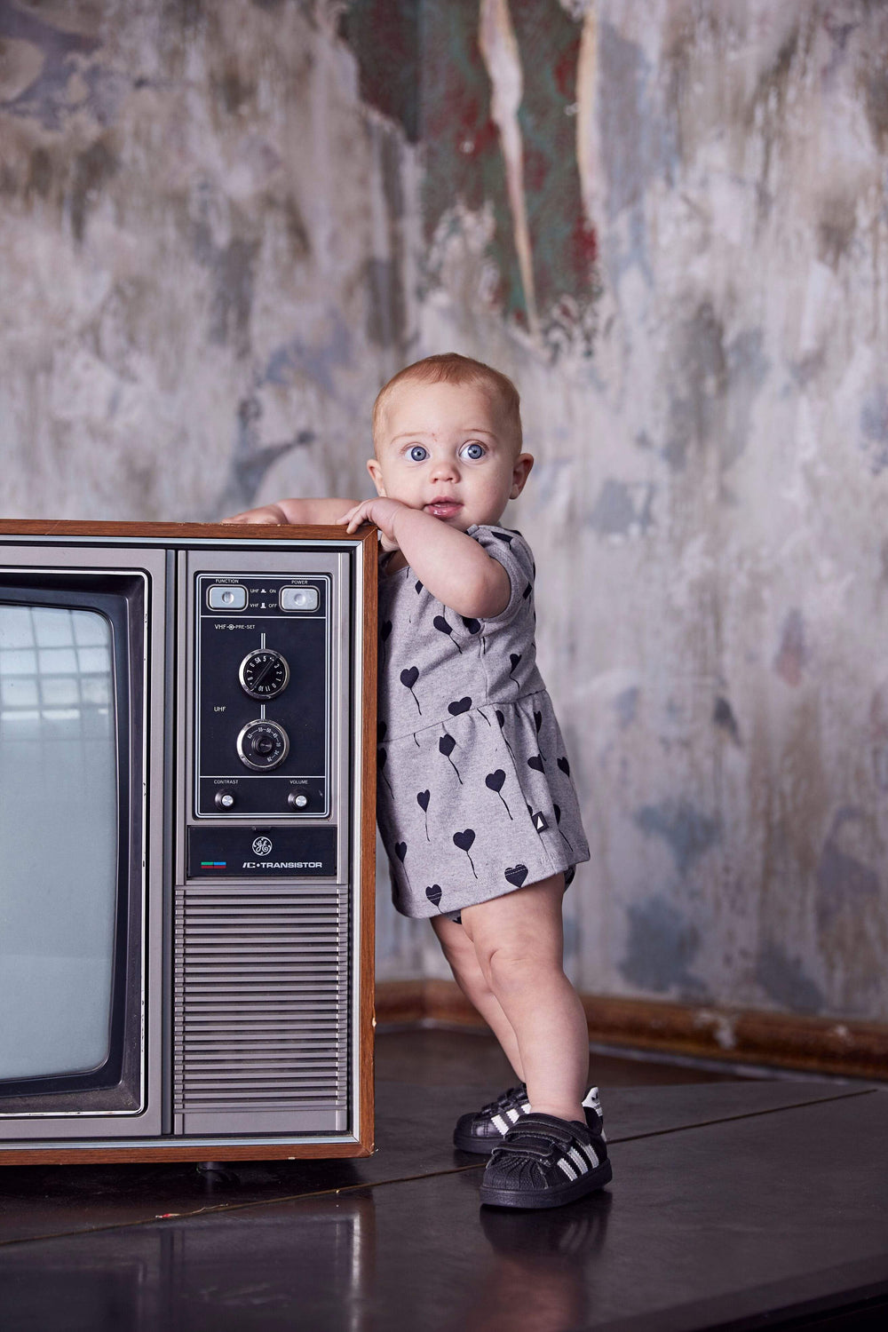 A baby is standing in front of an old tv wearing an Anarkid Organic Cotton Frill Onesie - LUCKY LAST - 0-3 MONTHS.
