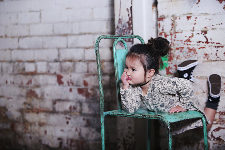 A little girl is sitting on a chair made of Anarkid Organic Cotton Moss Pit Band Swing Dress - LUCKY LAST - 0-3 MONTHS.