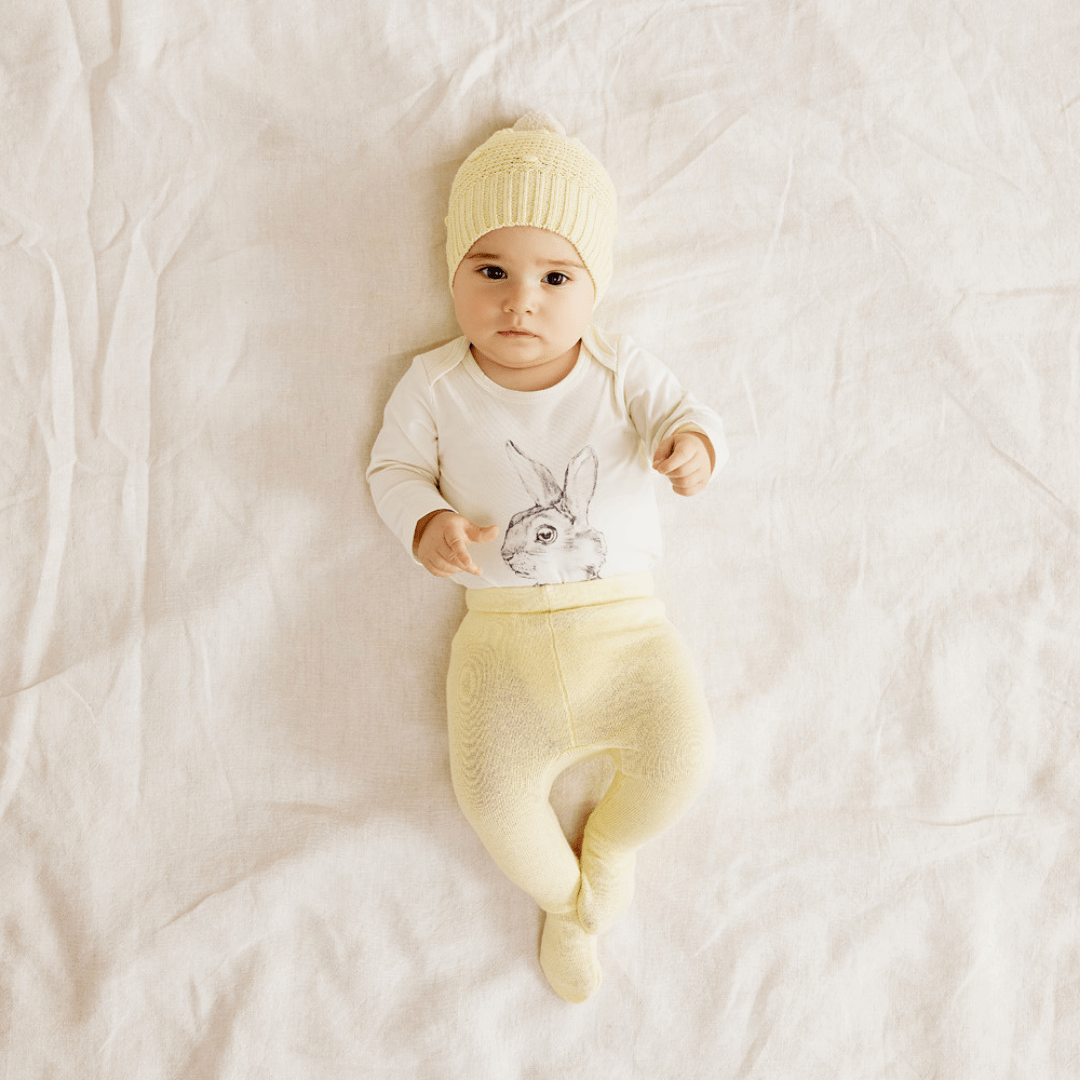 Baby-Lying-Down-Wearing-Wilson-and-Frenchy-Knitted-Leggings-with-Feet-Pastel-Yellow-with-Bunny-Onesie-Naked-Baby-Eco-Boutique