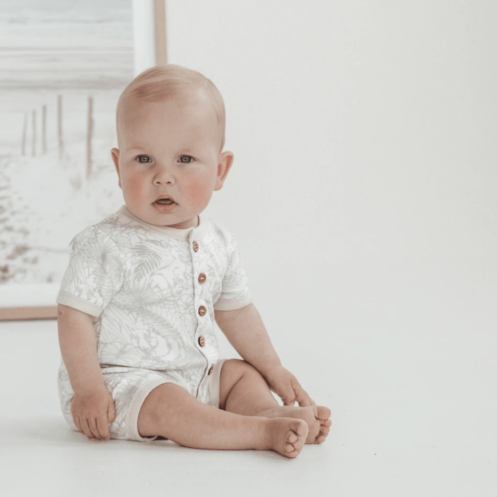 A baby wearing an Aster & Oak Organic Animal Button Romper - LUCKY LASTS - NEWBORN sitting on the floor in front of a picture.