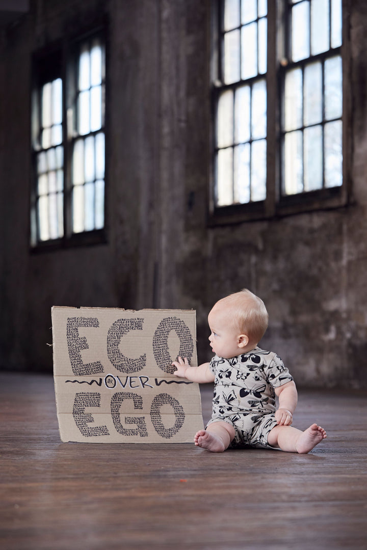 An eco-conscious baby sitting on the floor wearing an Anarkid Organic Cotton Short-Sleeve Romper with a sign that says eco-ego.