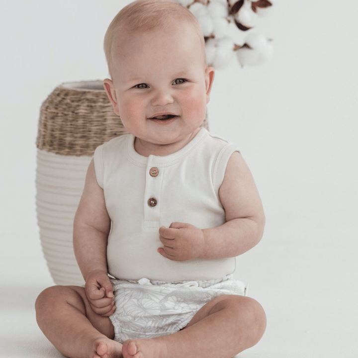 A baby is sitting on the floor in front of a potted plant wearing Aster & Oak Organic Animal Bloomers.