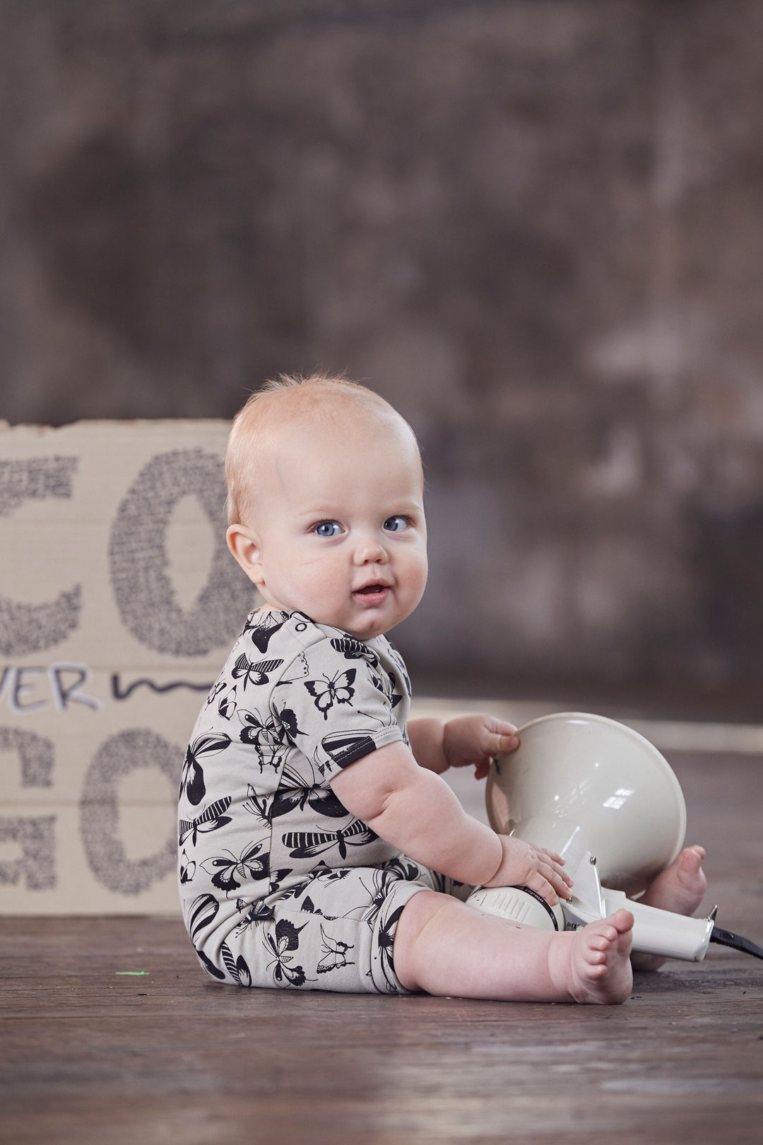 A baby sitting on a wooden floor wearing an Anarkid Organic Cotton Short Sleeve Romper with a GOTS certified organic cotton short sleeve romper with a sign on it.
