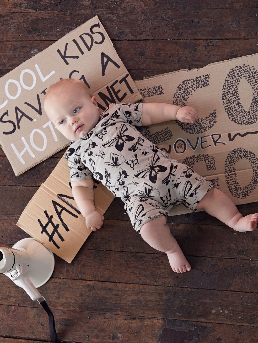 A baby laying on a wooden floor with Anarkid Organic Cotton Short-Sleeve Romper (Multiple Variants) and GOTS certified cardboard signs.