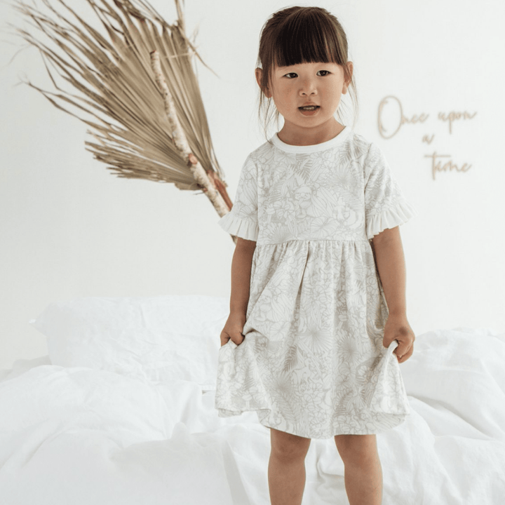 A little girl in an Aster & Oak Organic Animal Frill Skater Dress - LUCKY LAST - 2 YEARS, made of organic cotton, standing on a bed.