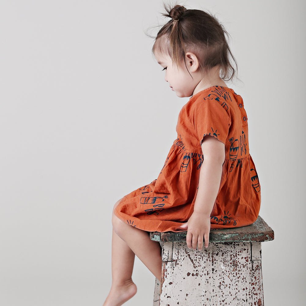 A little girl in an Anarkid Organic Cotton Beach Day Woven Dress - LUCKY LAST - 0-3 MONTHS ONLY sitting on a stool at the beach.
