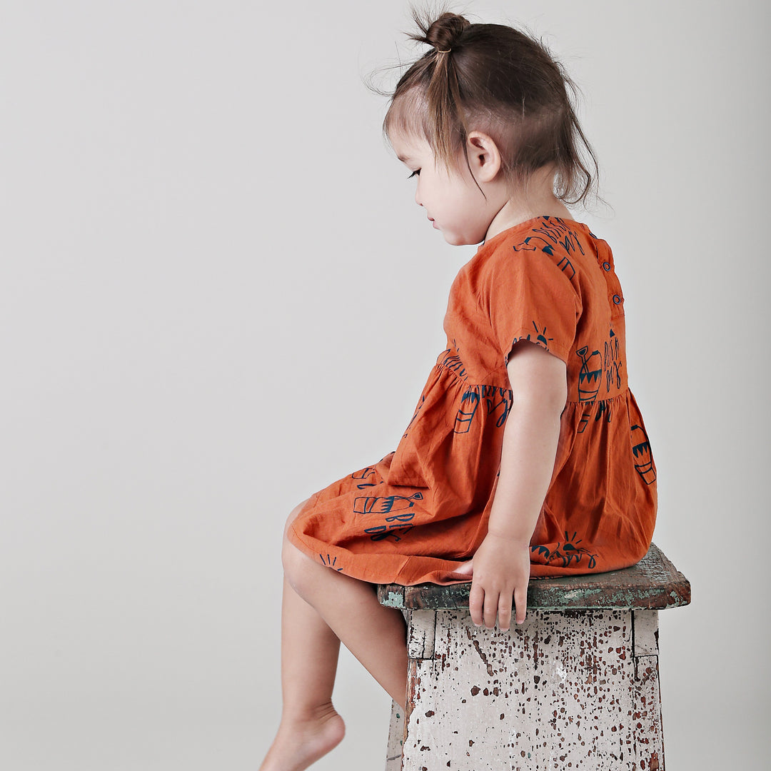 A little girl in an Anarkid Organic Cotton Beach Day Woven Dress - LUCKY LAST - 0-3 MONTHS ONLY sitting on a stool at the beach.