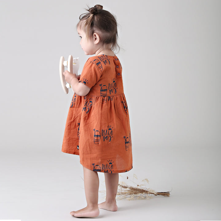 A little girl in an Anarkid Organic Cotton Beach Day Woven Dress - LUCKY LAST - 0-3 MONTHS ONLY holding a wooden toy on a beach day.