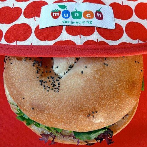 A sandwich with a poppy seed bun presented in a Munch Reusable Lunch Wrap by LUCKY LASTS, KIWI ONLY, set against a red background with white apple patterns.
