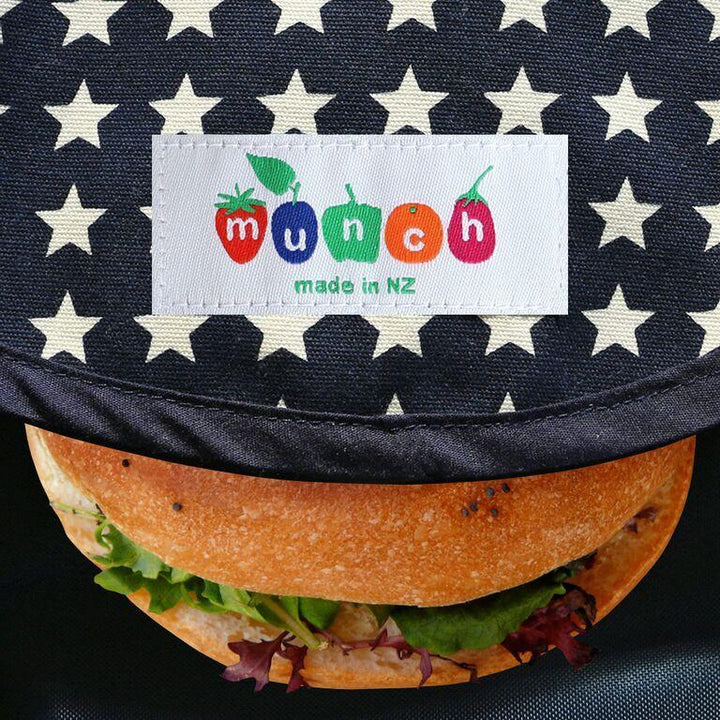 Burger with a label reading "Munch Reusable Lunch Wrap - LUCKY LASTS - KIWI ONLY" against a star-patterned background, wrapped in sustainable, reusable lunch wraps.